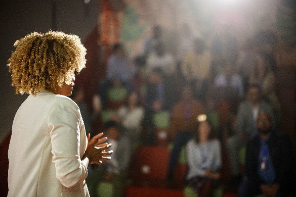 A lady in a taupe suit gives a presentation in an auditorium.